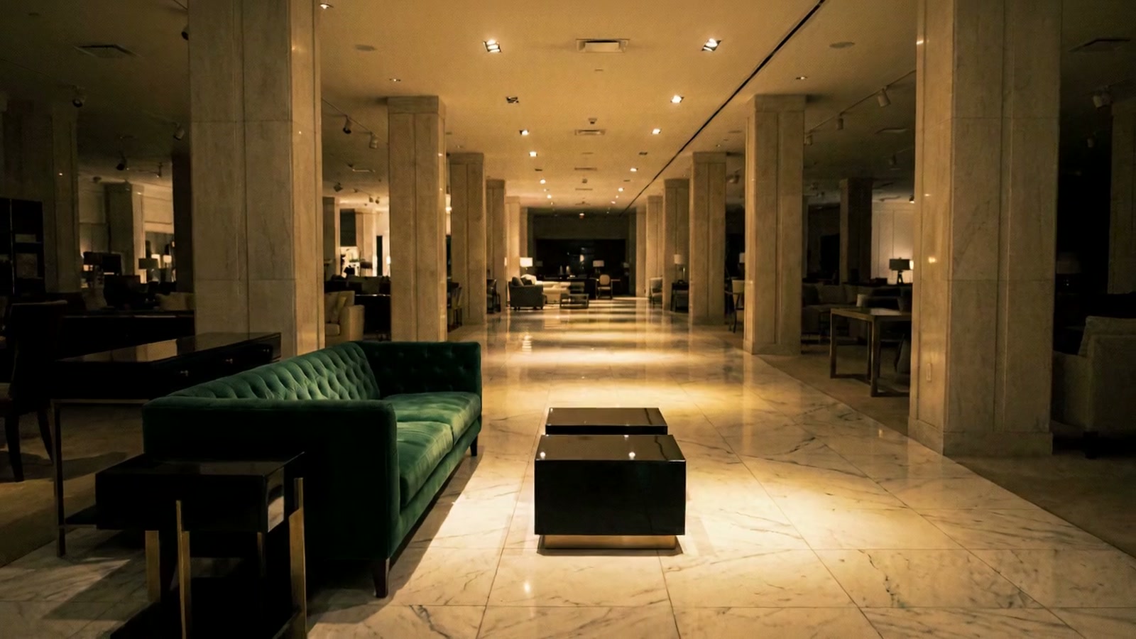 A marble department-store lounge lit low after closing, empty, with a green velvet sofa and a long corridor receding into the dark.